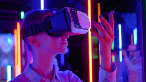 Slow motion: woman using virtual reality headset and looking around at interactive technology exhibition with colorful illumination. VR, futuristic, retrowave, immersive, entertainment concept