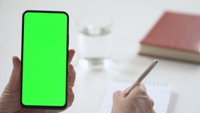 Close-up shot of green screen template smartphone in female hands at home offise, girl is watcing content, makes notes in a notebook. Modent technology and information concept.