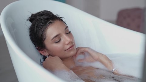 Close up relaxed woman touching skin with foam in bath. Top view of sexy girl taking bathtub at home. Romantic brunette woman relaxing bath in slow motion.