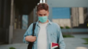 Attractive student man with green eyes in a mask look at camera hold books. Stand on street near university or office building. Coronavirus pandemic covid-19. Close up. Slow motion