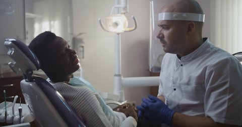 Adult dental man talking to black teen boy in chair and telling about new healthy treatment