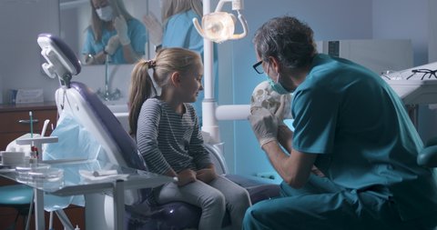 Professional kind dentist holding human skull model while showing teeth anatomy to little patient