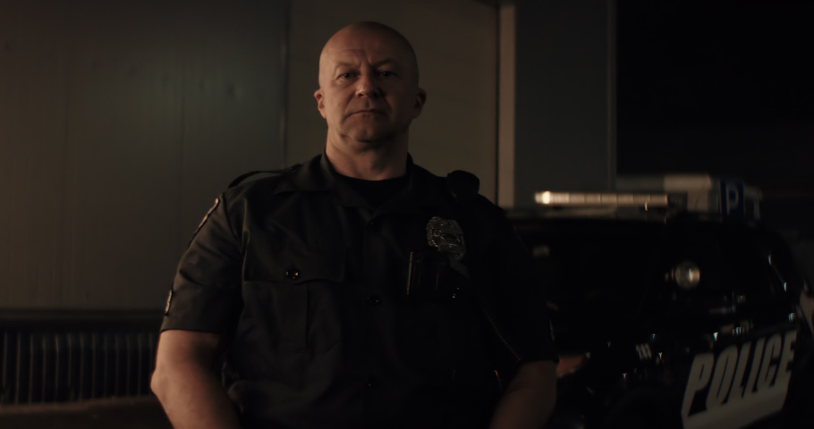 Caucasian male police officer posing against police car with flashing lights at night. Shot on RED cinema camera with 2x Anamorphic lens Royalty-Free Stock Footage #1061365864