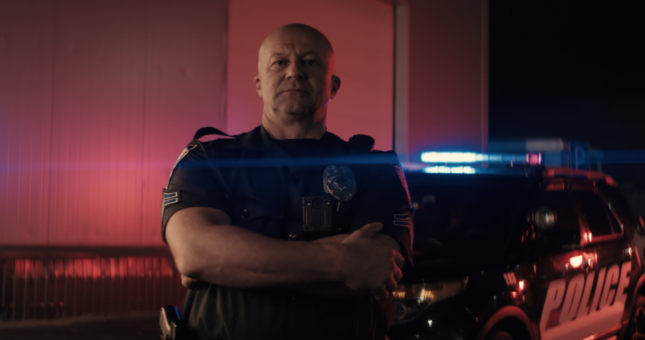 Caucasian male police officer posing against police car with flashing lights at night. Shot on RED cinema camera with 2x Anamorphic lens