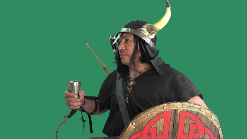 Portrait of formidable commoner warrior against green background, raises silver horn with mead and drinks, vino. Mature man in helmet with horns and with wooden shield
