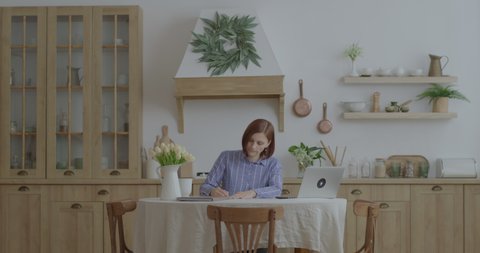Young 30s business woman working with paper documents and laptop at kitchen. Female entrepreneur working from home. Woman signing documents at home office.