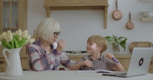 60s grandmother making online homework with preschool grandson at home. Senior woman in glasses and young boy studying online with laptop. Online education and distance learning process.