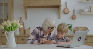 60s grandmother making online homework with preschool grandson at home. Senior woman in glasses and young boy studying online with laptop. Online education and distance learning process.