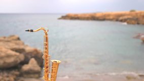 Blurry defocused beautiful golden alt tenor saxophone stands against backdrop of blurry sea waves. Music screensaver romance. Copy space for your text. Mini model real saxophone. Slow motion video.