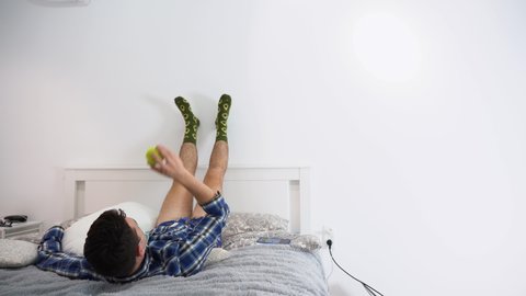 Bored adult male businessman wear plaid shirt, protective medical face mask, blue underpants, lies on gray bed, raised his legs against white wall background. Man plays with yellow ball. Copy space.
