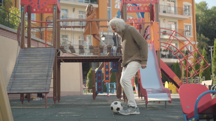 Wide shot of positive male retiree playing with football ball outdoors as children having fun at playground at the background. Portrait of carefree Caucasian senior man enjoying pension outdoors. Royalty-Free Stock Footage #1061381170