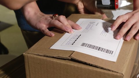 Close-up view of hands of women packing clothes and thank-you letter in box and attaching shipping label with adhesive tape while preparing order for customer of online fashion store