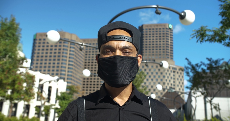 Portrait of young man wearing cap and black covid-19 protective face mask and looking upwards with faith against building outdoors