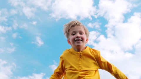 Happy child jumps very high on a trampoline against the blue sky. Blond boy jumps on a trampoline. Slow motion bottom view of a cheerful boy jumping on a trampoline. Children happiness. 