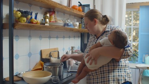 Woman housewife holding sleeping infant and cooking dinner in kitchen talking to older children. Babysitter rocking newborn and preparing meal for preschool kids in kitchen