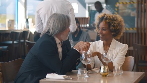 Diverse couple holding hands and talking on romantic date in restaurant. Portrait of mature caucasian boyfriend and young afro-american girlfriend celebrating anniversary in cafe