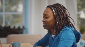 Young african woman using headset and laptop for online communication. Side view of smiling afro-american freelancer talking to client via video conference sitting at table in home office