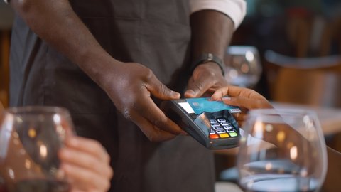 Close up customer make contactless credit card payment after eating in restaurant. African waiter holding terminal for client paying for dinner in cafe