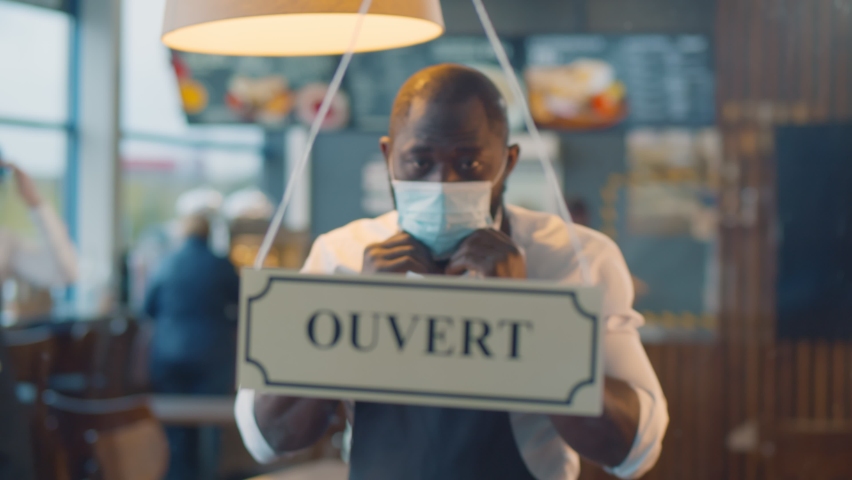 African waiter in safety mask turning ouvert ferme sign on french cafe glass door. Afro staff of restaurant turning open closed sign in french closing because of covid-19 epidemic Royalty-Free Stock Footage #1061388991