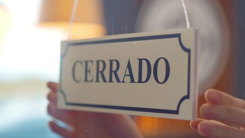 Close up of human turning abierto cerrado sign hanging on glass door of spanish cafe. Owner or seller turning open closed sign written in spanish on window of store