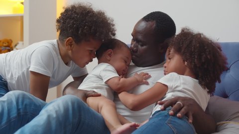 Happy african american single father with little children lying on bed. Portrait of young afro man holding newborn baby with older son and daughter lying together in bedroom