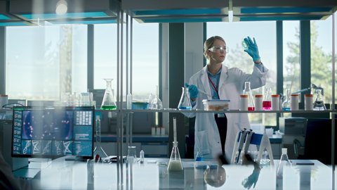 Beautiful Female Medical Scientist Wearing White Coat and Safety Glasses uses Micropipette while Examining Testing Sample. Innovative, Experimental Drugs Research, Development in High-Tech Laboratory