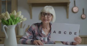 Senior teacher in glasses showing sheet of paper with alphabet letters looking at camera. Online education and distance learning process. Webcam view of working from home woman talking.