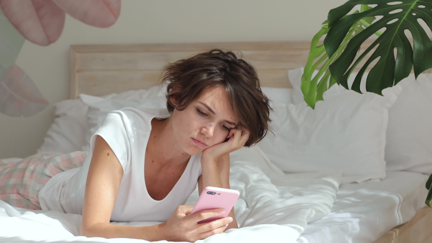 Sad upset dissatisfied boring young woman in white t-shirt pajamas home wear lying in bed spending time in bedroom hold mobile cell phone say no i do not need it put aside. Bad mood lifestyle concept Royalty-Free Stock Footage #1061397046