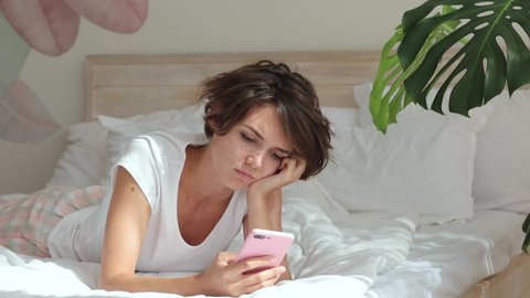 Sad upset dissatisfied boring young woman in white t-shirt pajamas home wear lying in bed spending time in bedroom hold mobile cell phone say no i do not need it put aside. Bad mood lifestyle concept
