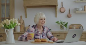 Senior woman talking online by laptop sitting at wooden kitchen. Smiling elderly woman having online conversation. Granny stays at home and chats with relatives online. 70s woman drinks orange juice.