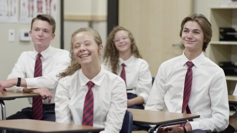 Happy secondary school students sit in classroom in uniforms, boy raises hand