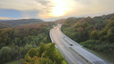 Aerial view of traffic highway through a green forest tree during colorful sunset autumn season. Green energy eco electric car no pollution.
