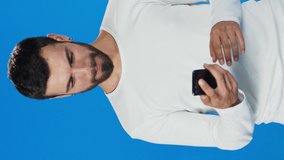 The teenager laughing at what he saw on the mobile phone. Handsome bearded young man 20s in jeans shirt white t-shirt isolated on blue background in studio.Video for the vertical story.