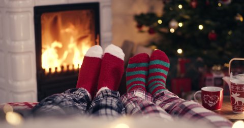 Couple feet in cozy christmas woolen socks near fireplace with decorated xmas tree and tee cup in background shot in 4k