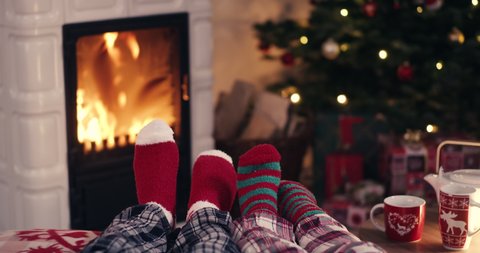 Couple feet in cozy christmas woolen socks near fireplace with decorated xmas tree and tee cup in background shot in 4k