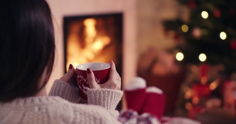 Over the shoulder young woman relaxing near fireplace with cup of tee in cozy christmas woolen socks with decorated xmas tree in background shot in 4k