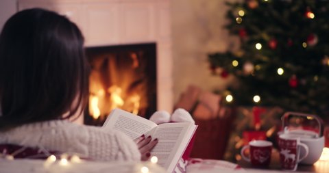 Over the shoulder young woman relaxing near fireplace reading a book in cozy christmas woolen socks with decorated xmas tree in background shot in 4k