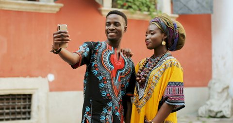 African young stylish and attractive couple in traditional outfits standing together outside and posing to the smartphone camera while taking selfie photo.