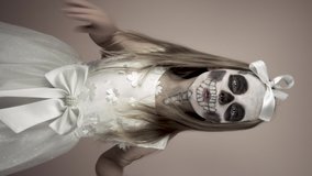 A little girl in a beautiful white dress with terrible make-up on her face scares. Scary skull on her face, she scares and grimaces. Halloween costume. Face painting.Vertical video.