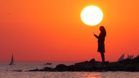 Woman silhouette at sea shore, big sun disk on background. Cool autumn weather, tourist lady take pictures and videos using smartphone, stand at waterside in beautiful sunset time