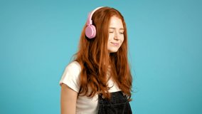Pretty young girl with red hair listening to music, smiling, dancing in pink headphones in studio against blue background. Music, dance, radio concept, slow motion.