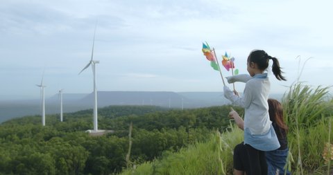Asian mother and daughter are playing with turbine toy at the wind turbine field with fun together. Child girl and mother enjoy with beautiful scenery view of nature in slow motion shot.