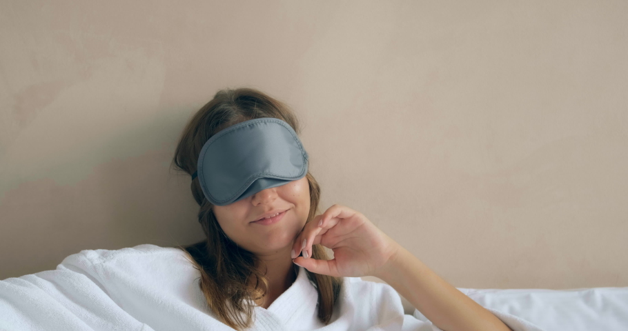 smiling woman in white bathrobe takes off black sleep mask sitting against wall in hotel room in morning closeup