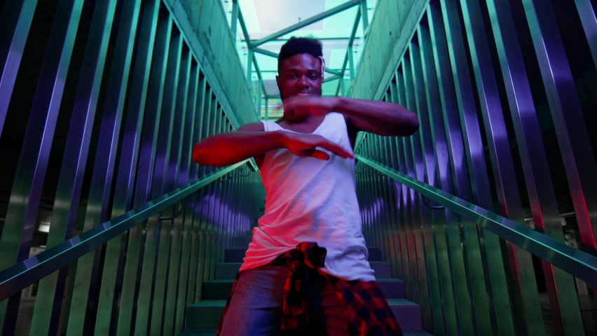 Young Black Guy Dancing On The Stairs. He Has Stylish Hairstyle And Clothes Tied At The Waist. He Moves Rhythmically. Lighting Creates Blue And Green Colors. Royalty-Free Stock Footage #1061421046