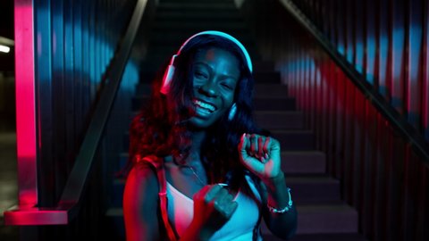Portrait Of Black Young Girl In Headphones. She Stands On The Background Of The Stairs. Lighting In Different Colors. Pretty Girl With Long Hair. She Smiles And Dances.