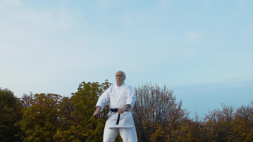 Against the background of the sky and trees, an old man athlete trains formal karate exercises