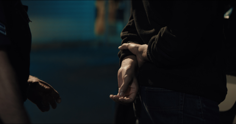 CLOSE UP Police officer handcuffs a suspect near police car, African-American Black criminal. Lights flashing in the background. Shot with RED cinema camera and 2x Anamorphic lens Royalty-Free Stock Footage #1061427715