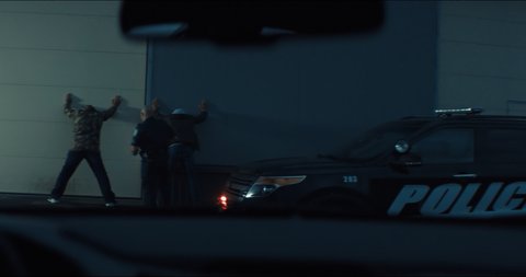 WIDE Police officer searches two suspects in the street at night. Police car lights flashing in the background. Shot with RED cinema camera and 2x Anamorphic lens