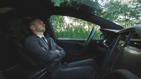 Businessman sleeping behind self-driving steering wheel in autonomous autopilot driverless electric car. Deeply asleep man driving along the countryside road in luxury all-electric vehicle