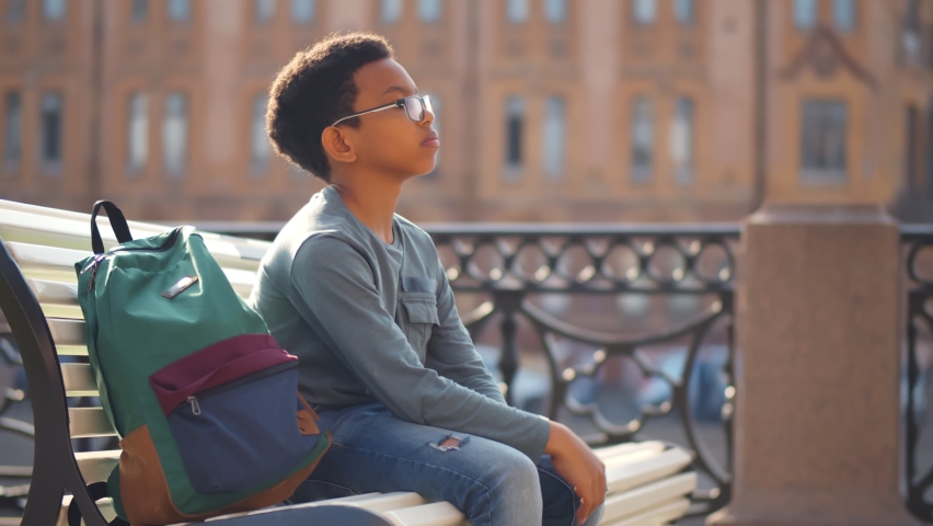 Side view of sad mixed-race schoolboy sitting alone on bench outdoors. Upset and worried african preteen boy with backpack sitting on street bench anxious about exam Royalty-Free Stock Footage #1061428285